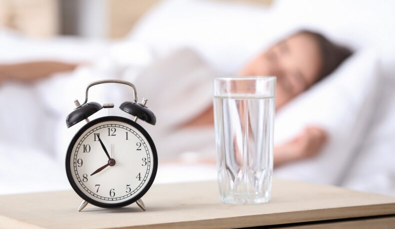Detaliu cu pahar cu apă lângă ceas și fată care doarme în fundal, sugestiv pentru de ce e bine să bei apă dimineața, la trezire