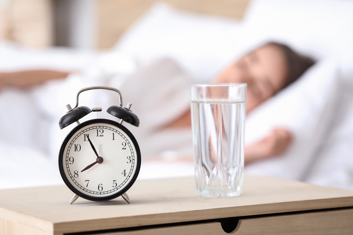 Detaliu cu pahar cu apă lângă ceas și fată care doarme în fundal, sugestiv pentru de ce e bine să bei apă dimineața, la trezire
