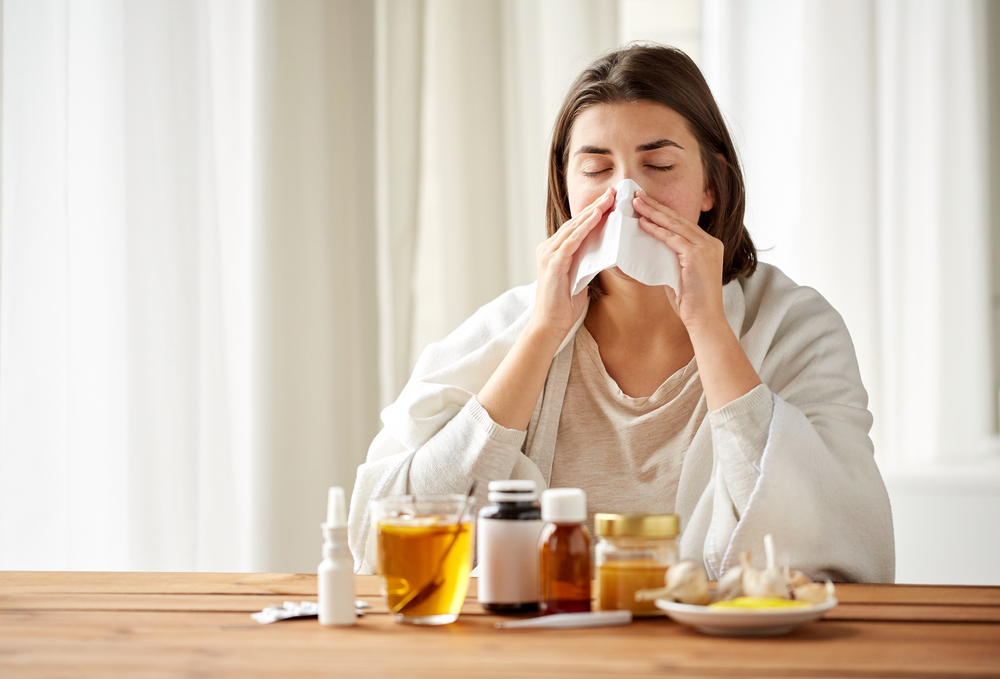 O femeie tânără ține un șervețel la nas, în timp ce stă la o masă cu remedii naturale pentru răceală