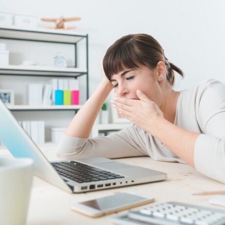 Femeie obosită la birou, care cască în fața laptopului