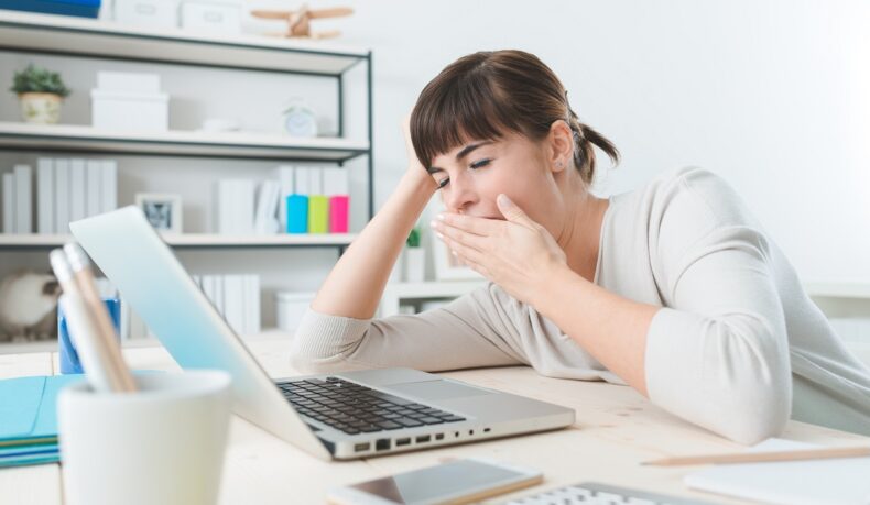 Femeie obosită la birou, care cască în fața laptopului