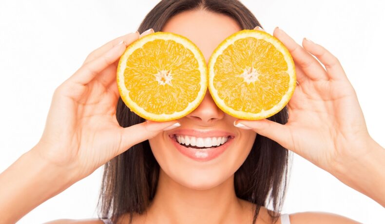 femeie cu două felii de portocală la ochi, așezate ca ochelarii, citricele fiind unul dintre alimentele care conțin vitamine pentru ochi