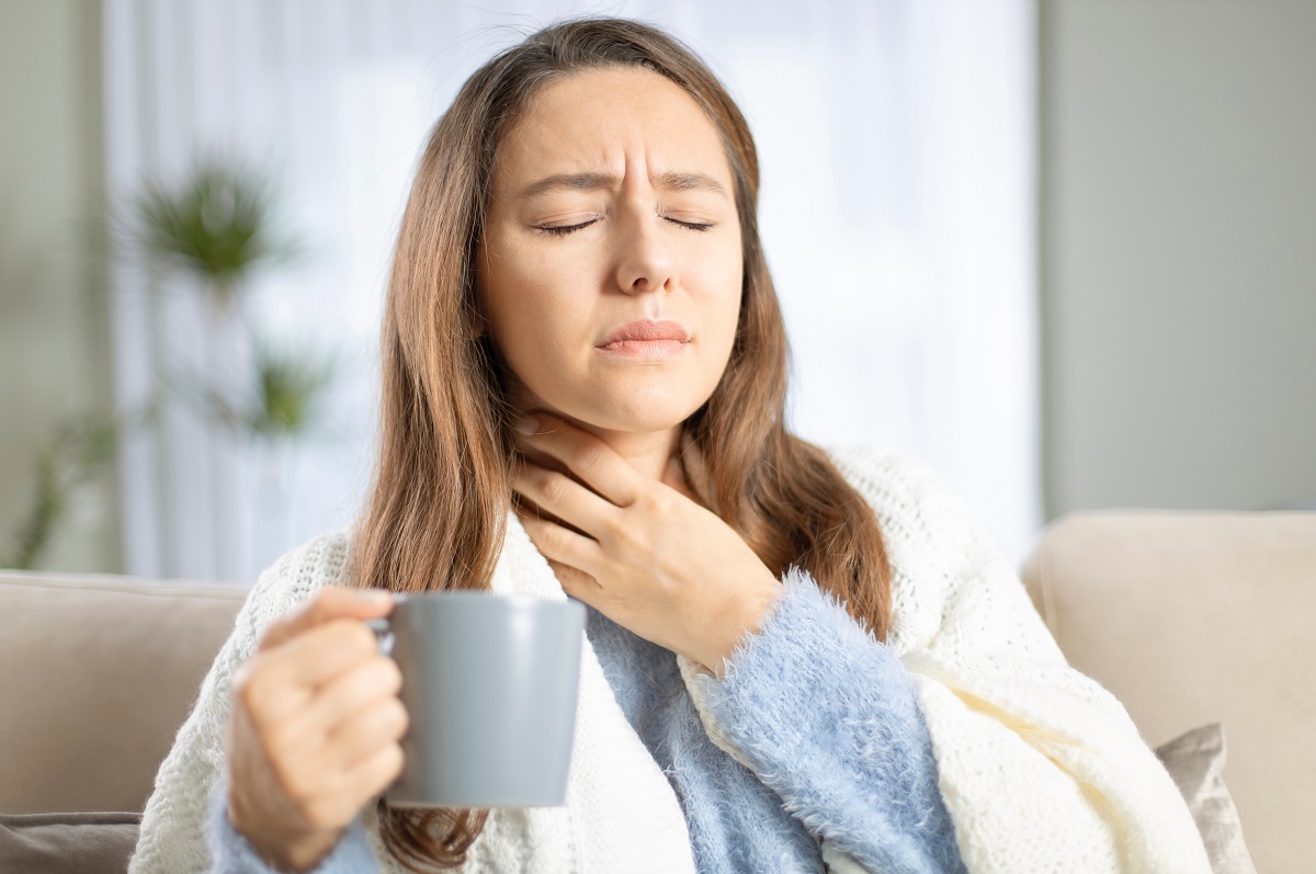 Femeie cu durere în gât și cană de ceai în mână, sugestiv pentru remedii pentru durerea în gât