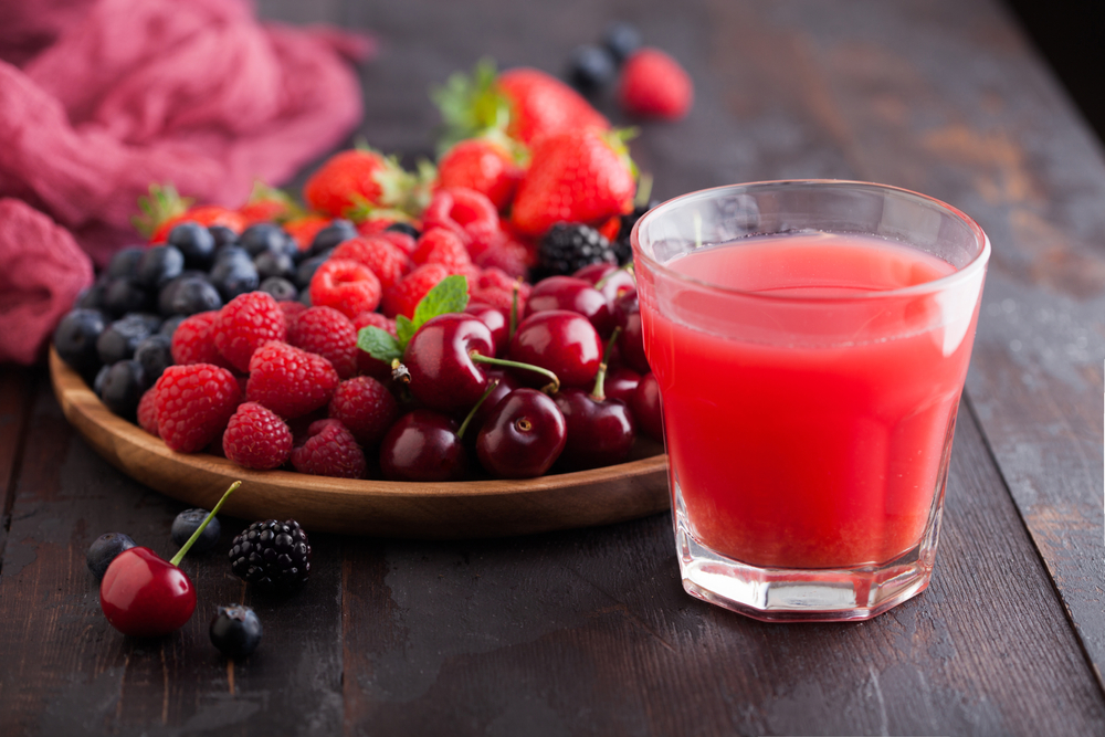 Un pahar cu suc de fructe, lângă o farfurie cu zmeură, coacăze, cireșe și mure