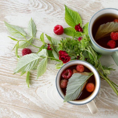 Două căni cu ceai de zmeură roșie, fructe de zmeură și frunze