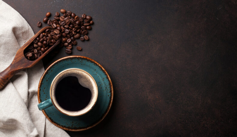 O ceașcă cu cafea, lângă o lingură cu boabe de cafea, puse pe un șervet