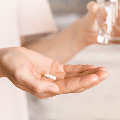Detaliu cu mână care ține o pastilă albă și un pahar cu apă, sugestiv pentru vitamine care previn scăderea masei musculare