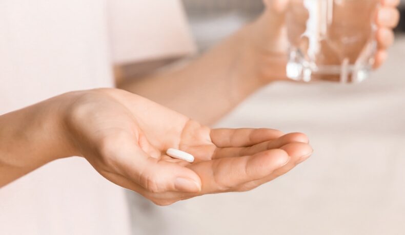 Detaliu cu mână care ține o pastilă albă și un pahar cu apă, sugestiv pentru vitamine care previn scăderea masei musculare