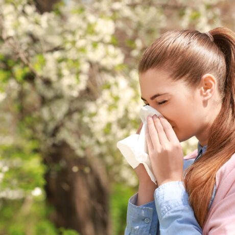 Fată care își suflă nasul în aer liber, lângă un copac înflorit, sugestiv pentru alergiile de primăvară