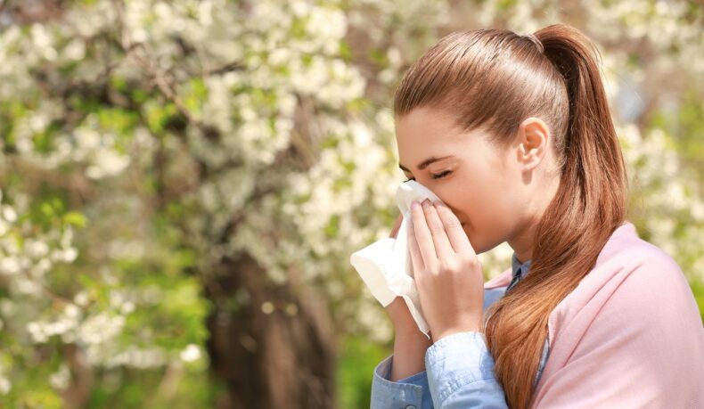 Fată care își suflă nasul în aer liber, lângă un copac înflorit, sugestiv pentru alergiile de primăvară