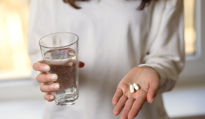 Femeie cu două pastile albe în palmă și cu pahar de apă, asemănătoare cu suplimentele cu magneziu și zinc