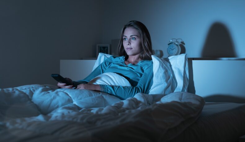 Femeie în par care se uită la televizor și ține o telecomandă în mână, sugestiv pentru obiceiul de a dormi cu televizorul aprins