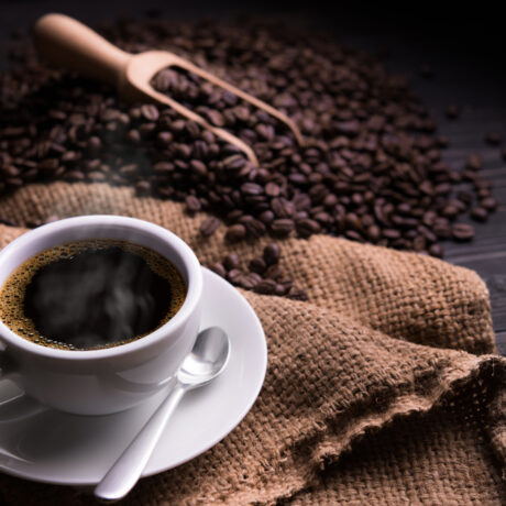 O ceașcă cu cafea pusă pe un șervet, lângă boabe de cafea și o lingură