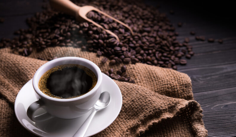 O ceașcă cu cafea pusă pe un șervet, lângă boabe de cafea și o lingură