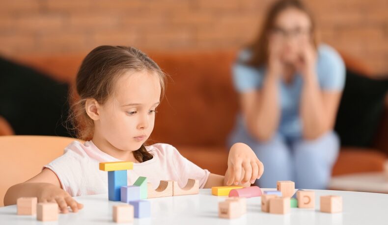 Fetiță care construiește dn cuburi de lemn la masă și mamă care o supraveghează în fundal, sugestiv pentru autismul la fete