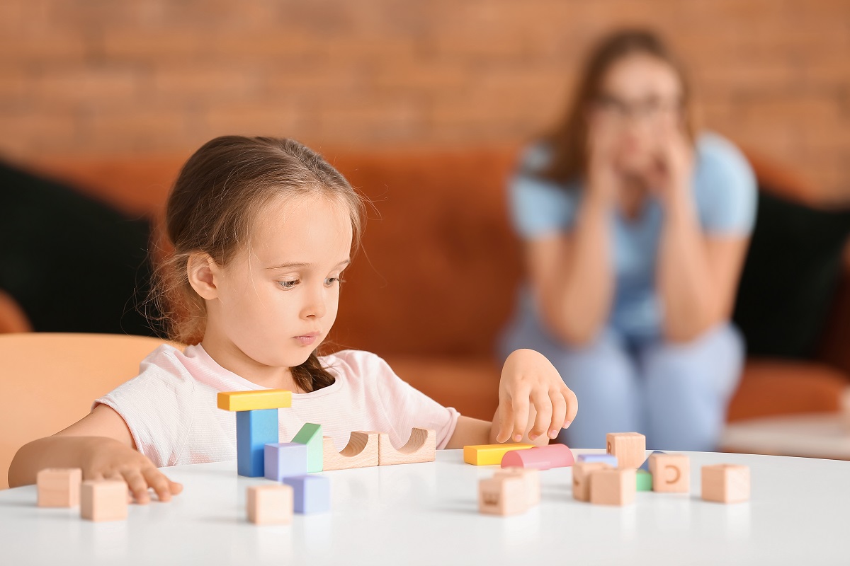 Fetiță care construiește dn cuburi de lemn la masă și mamă care o supraveghează în fundal, sugestiv pentru autismul la fete