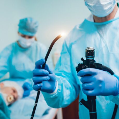 Medic cu endoscop în mână în prim plan și pacientă întinsă pe pat în fundal, alături de o asistentă care efectuează pregătirea pentru endoscopie