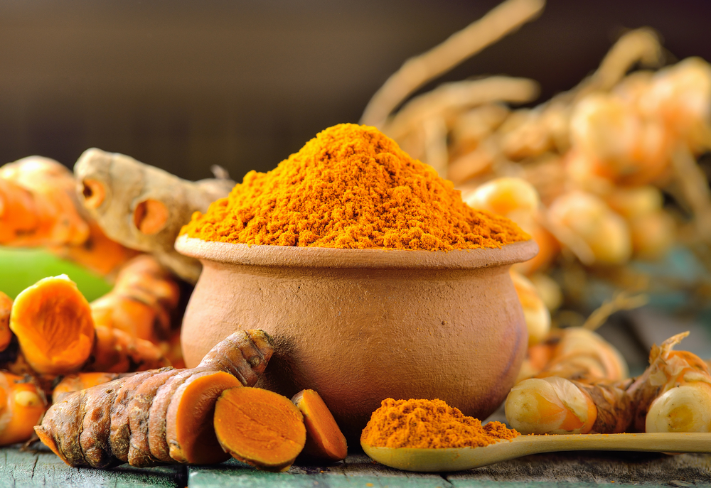 Un vas și o lingură cu pudră de turmeric, lângă rădăcini de turmeric