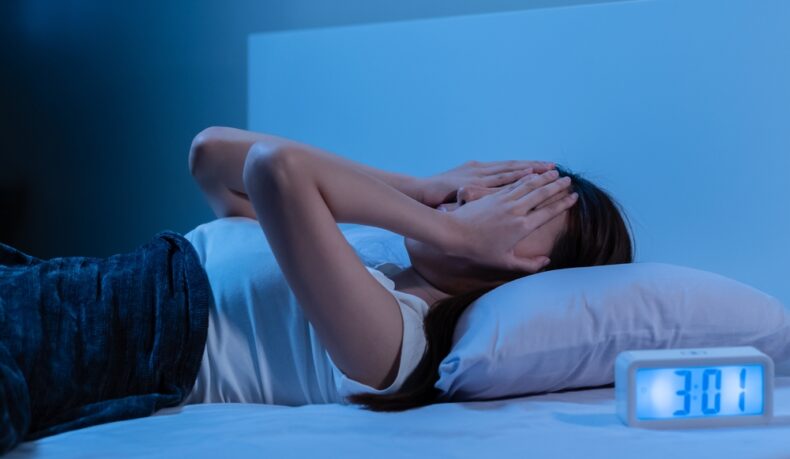 O femeie tânără stă întinsă în pat cu mâinile pe față lângă un ceas digital