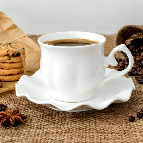 O ceașcă cu cafea pusă pe o farfurie, biscuiți, un sac cu boabe de cafea și anason