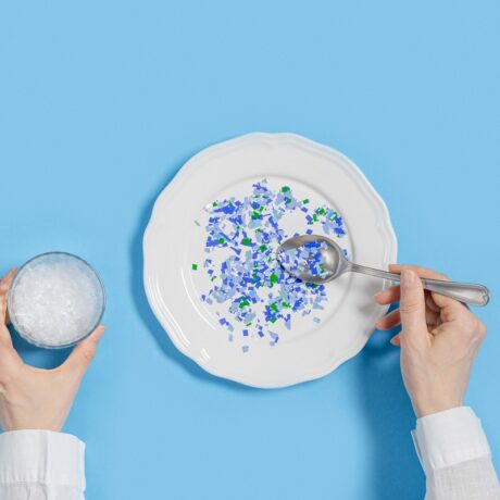 Detaliu cu persoană care mănâncă dintr-o farfurie cu bucăți mici de plastic colorat și ține un pahar cu plastic în interior, sugestiv petru alimentele care conțin cel mai mult plastic