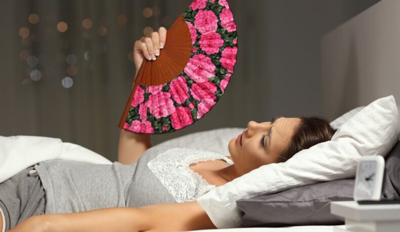 Femeie întinsă în pat cu evantai, sugestiv pentru transpirațiile nocturne