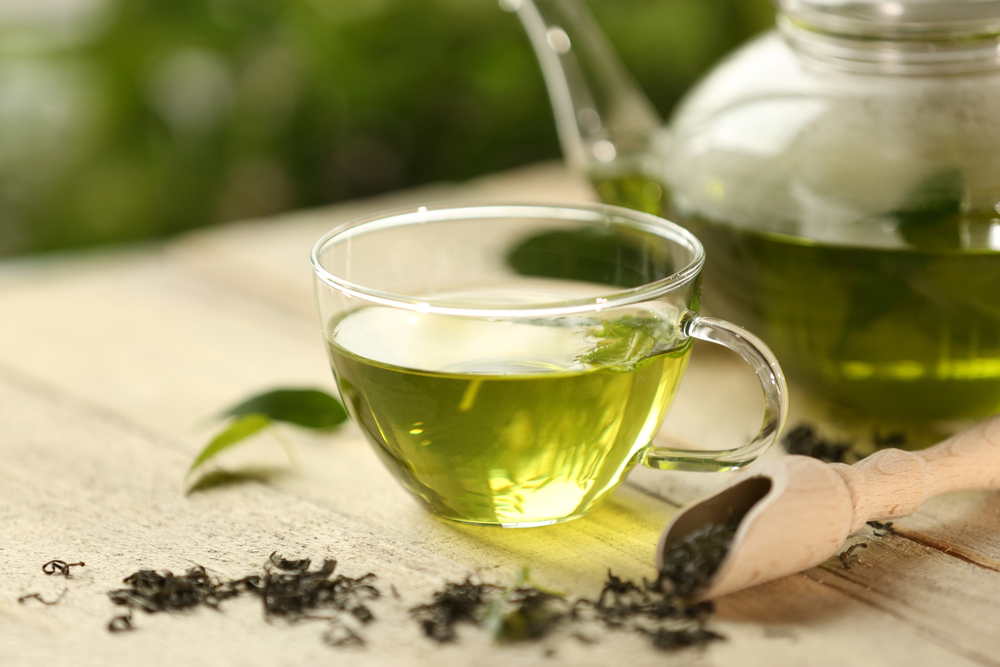 O ceașcă cu ceai verde, lângă un ceainic cu ceai verde și o lingură cu frunze uscate