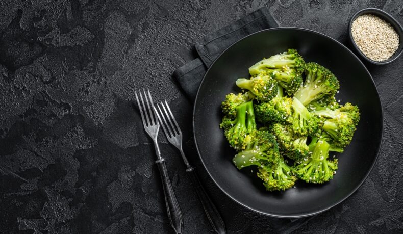 O farfurie cu broccoli fiert, pusă pe un șervet, lângă un castron cu susan și două furculițe