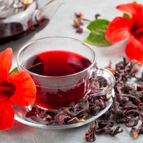 O ceașcă cu ceai de hibiscus, pusă pe o farfurie cu frunze uscate de hibiscus, alături de două flori de hibiscus și un ceainic