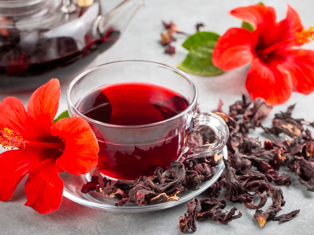 O ceașcă cu ceai de hibiscus, pusă pe o farfurie cu frunze uscate de hibiscus, alături de două flori de hibiscus și un ceainic