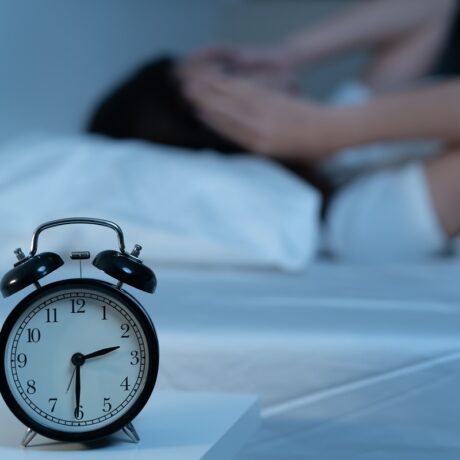 ceas în prim plan și femeie care stă în pat în fundal, sugestiv pentru insomnie