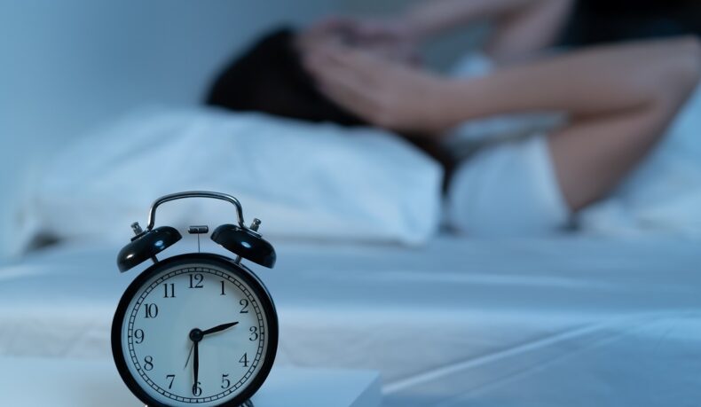 ceas în prim plan și femeie care stă în pat în fundal, sugestiv pentru insomnie