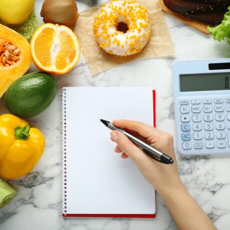 Detaliu mână de femeie care calculează caloriile și cu alimente în jurul ei, sugestiv pentru calculul deficitului caloric