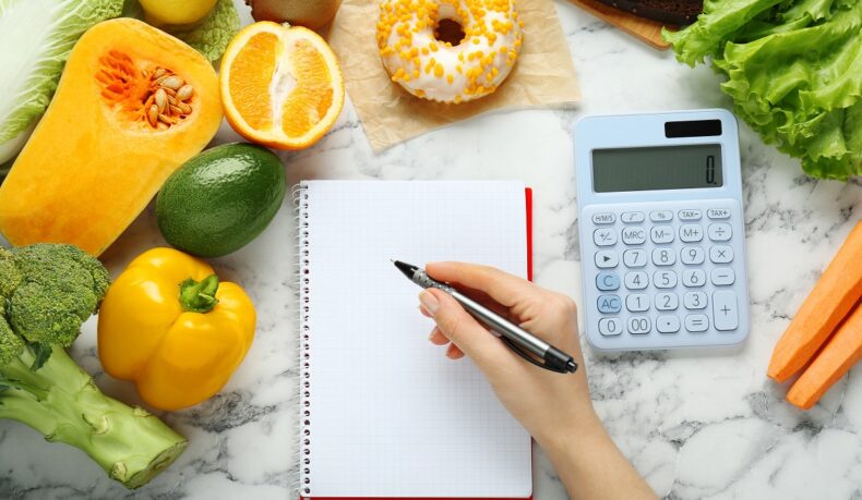 Detaliu mână de femeie care calculează caloriile și cu alimente în jurul ei, sugestiv pentru calculul deficitului caloric