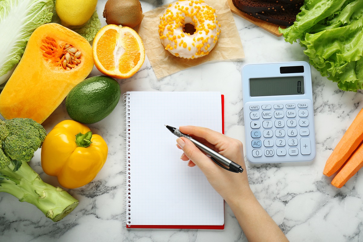 Detaliu mână de femeie care calculează caloriile și cu alimente în jurul ei, sugestiv pentru calculul deficitului caloric