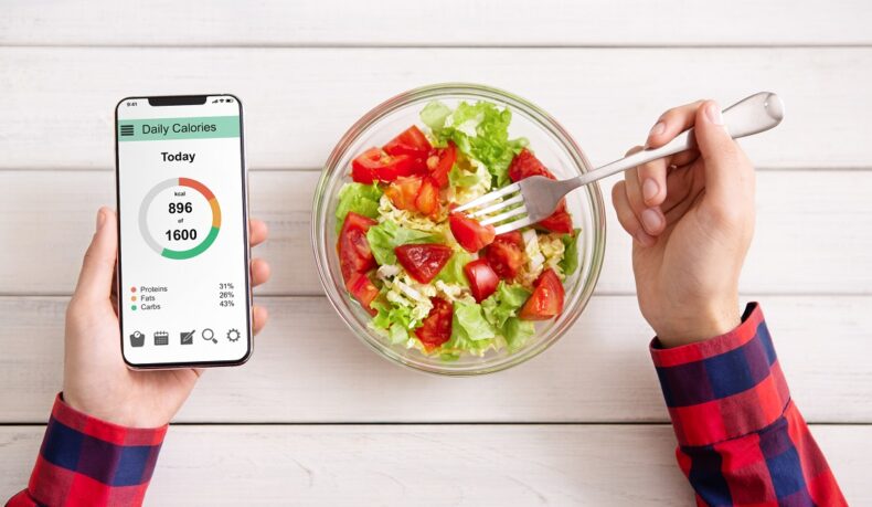detaliu cu un bol de salată și mâini care țin un telefon pe care se afișează aportul caloric, sugestiv pentru dietele cu deficit caloric