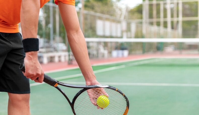 Un bărbat tânăr care ține o paletă de tenis în mână și o minge, pe un teren de tenis