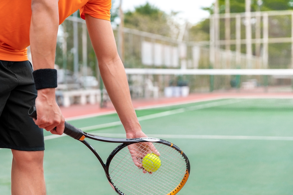 Un bărbat tânăr care ține o paletă de tenis în mână și o minge, pe un teren de tenis