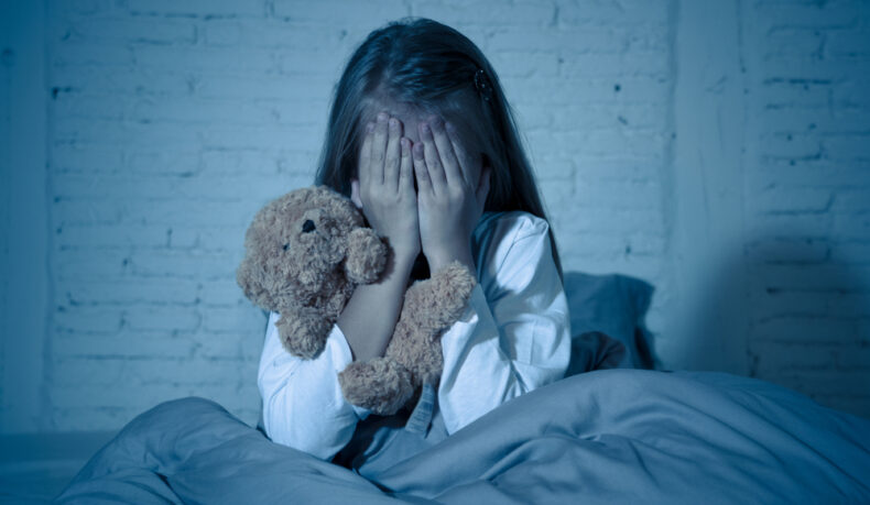 O fetiță care stă în pat cu un ursuleț de pluș și își ține mâinile pe față noaptea
