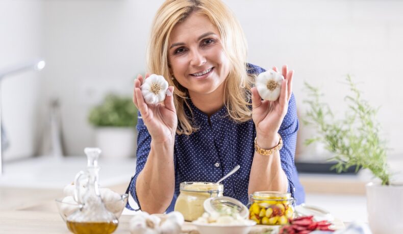 O femeie care ține în mâini două căpățâni de usturoi și are pe masă o pastă de usturoi și alte preparate din usturoi