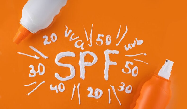 Sticle de plastic cu cremă de plajă, alături de cuvântul SPF scris cu cremă albă pe fundal portocaliu: SPF 50, 40, 30, 20