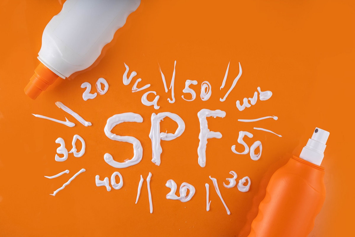 Sticle de plastic cu cremă de plajă, alături de cuvântul SPF scris cu cremă albă pe fundal portocaliu: SPF 50, 40, 30, 20