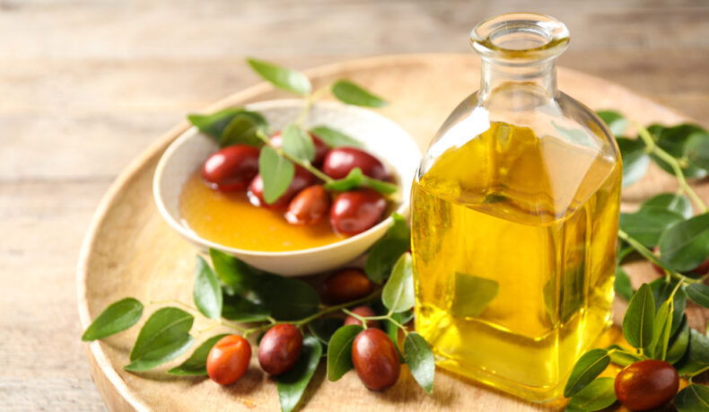 O sticlă cu ulei de jojoba, lângă o farfurie cu fructe și frunze de jojoba, puse pe un blat din lemn