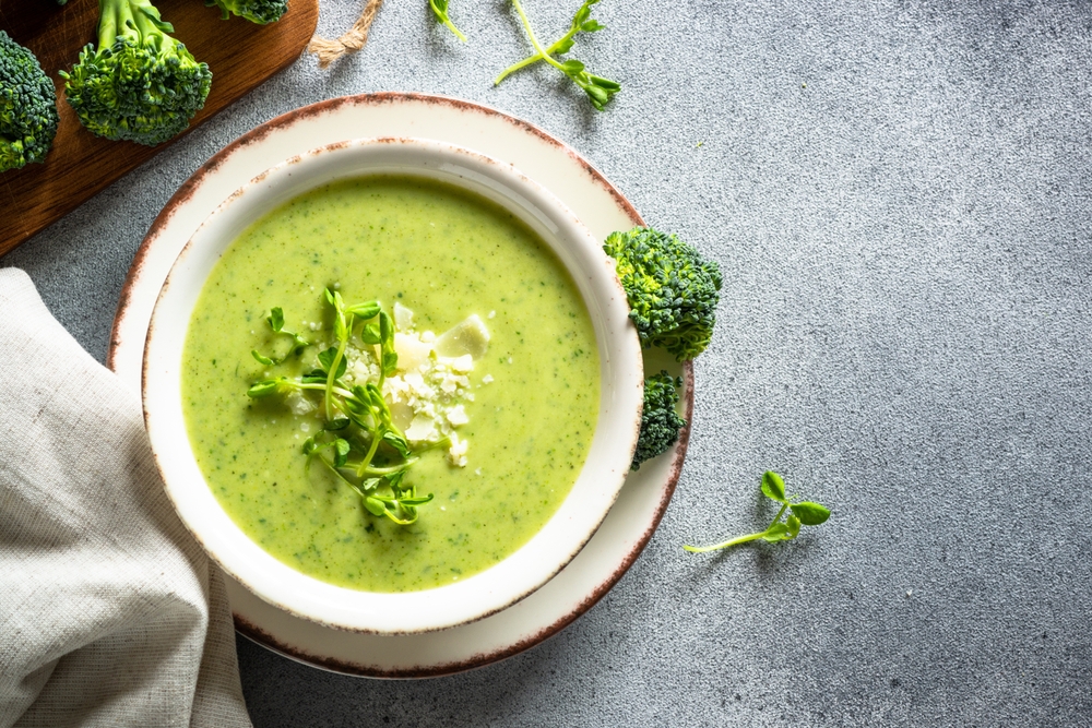 Un castron cu supă cremă de broccoli cu parmezan, pus pe o farfurie, lângă un tocător de lemn acoperit de broccoli și un șervet