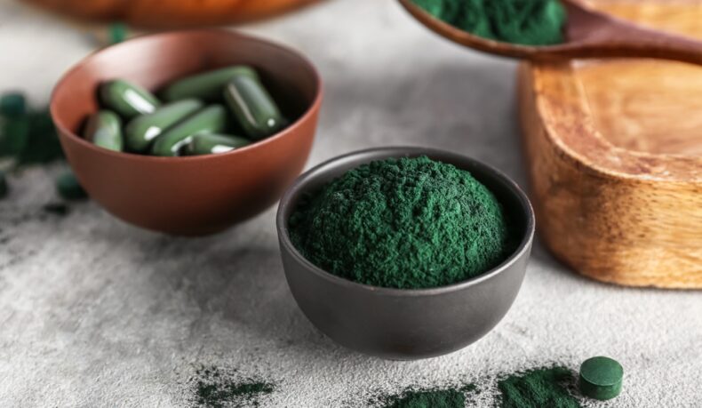 Un castron cu pudră de spirulină, un castron cu pastile de spirulină și o lingură cu pudră de spirulină
