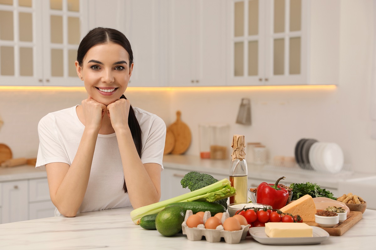 O femeie într-o bucătărie cu multe alimente pe masă: legume, ouă, unt și brânză