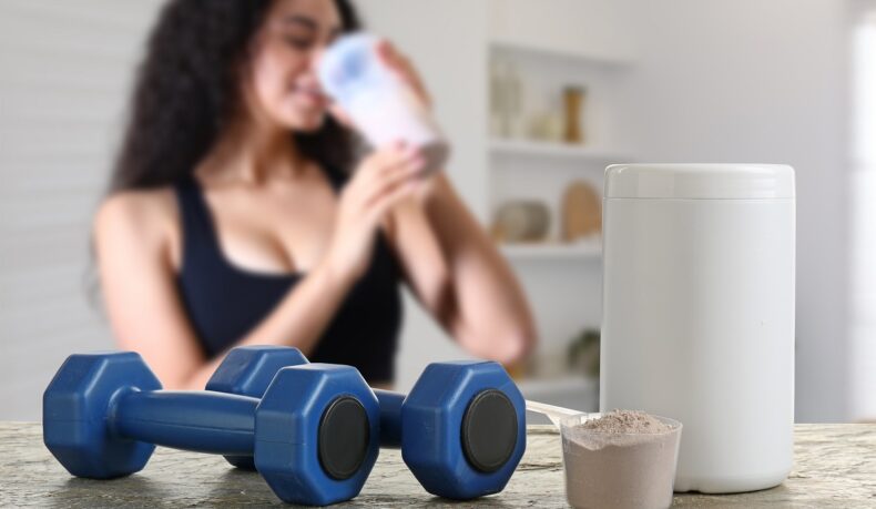 pudră de proteine sau de creatină și gantere în prim plan, cu femeie care bea un shake pe fundal